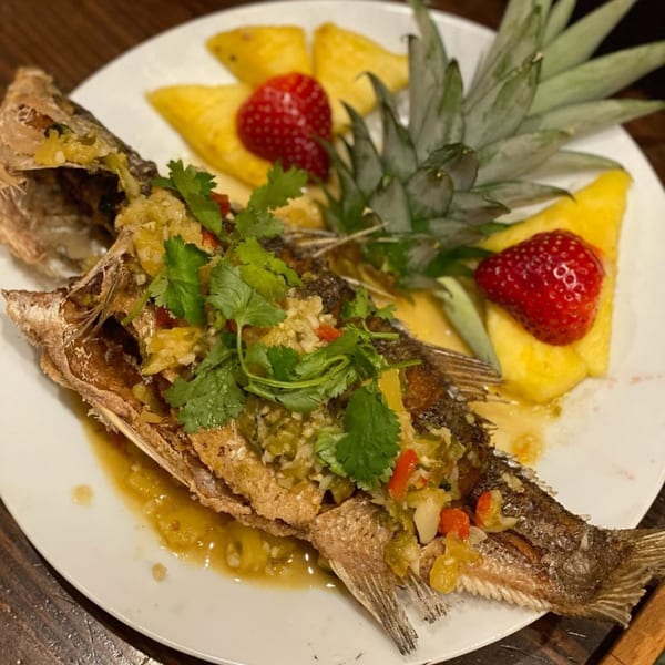 Whole fried fish with green chili sauce, cilantro, pineapple slices, and strawberries on a white plate.