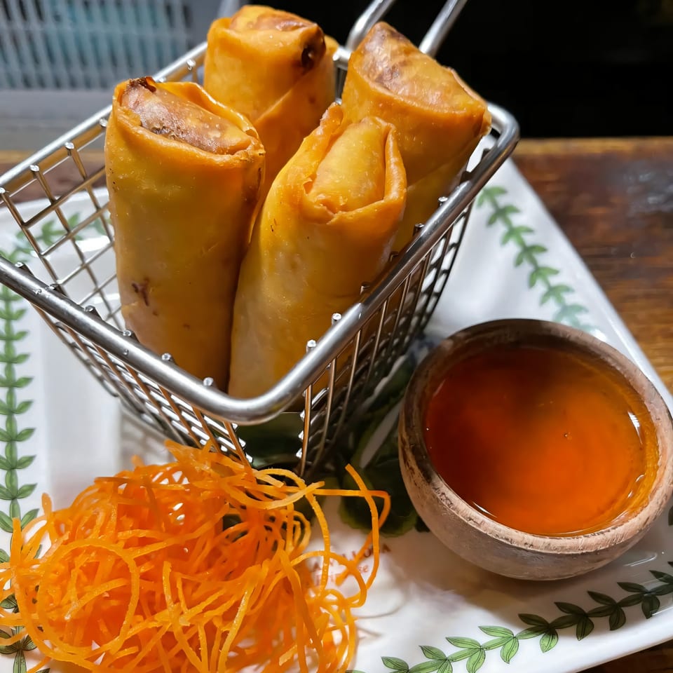 Crispy Thai spring rolls in a metal basket with shredded carrots and sweet chili dipping sauce.